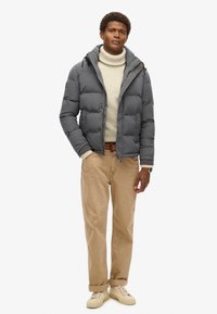 Szary puchowy kurtka z kapturem, zapinana na zamek, z elastycznymi mankietami, noszona na beżowym golfie, w połączeniu z khaki spodniami i beżowymi trampkami.