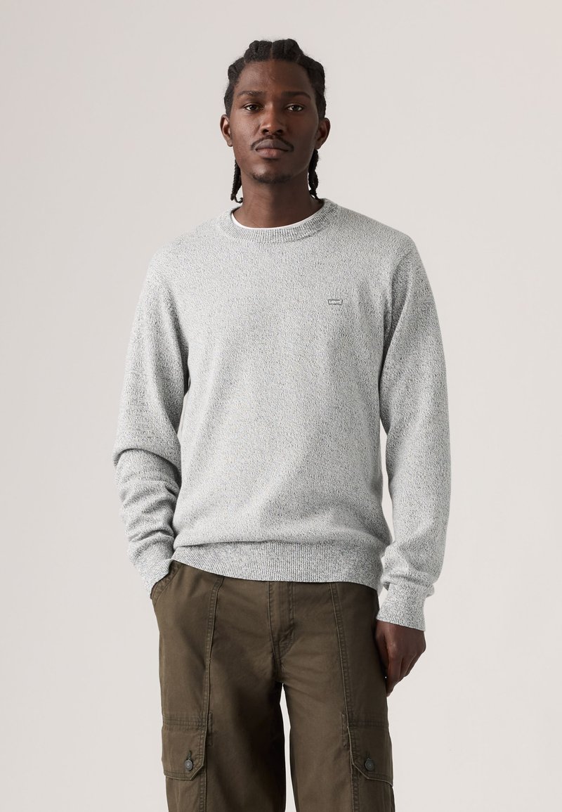 Mann mit geflochtenem Haar trägt einen hellgrauen Strickpullover und olivgrüne Cargohosen, steht mit einer Hand in der Tasche.