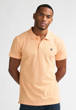 Hombre con una camisa polo de manga corta de color naranja claro con dos botones, de pie con las manos detrás de la espalda contra un fondo blanco liso.