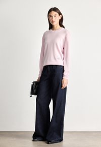 Ανοιχτό ροζ πουλόβερ με μακριά μανίκια και ριγωτές μανσέτες, συνδυασμένο με φαρδιά σκούρα μπλε τζιν. Μαύρη τσάντα, ελάχιστη επωνυμία στο πουλόβερ.