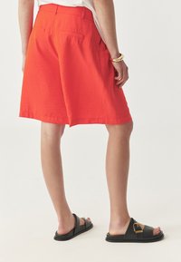 Rode shorts met een gestructureerde afwerking, middellange snit, voorzien van twee achterzakken. Gekleed met zwarte sandalen met een gouden gespdetail.