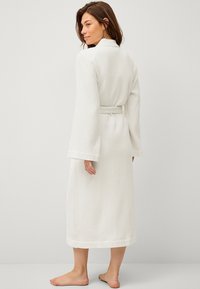 Robe en coton texturé blanc avec des manches longues, un col et un lien à la taille, présentant un motif en quadrillage et une longueur mi-mollet.