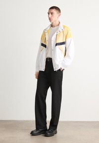 Jachetă color-block în alb, galben și bleumarin, având un închizător cu fermoar frontal și guler în picioare. Purtați-o peste o cămașă albă și pantaloni negri, cu pantofi negri masivi.