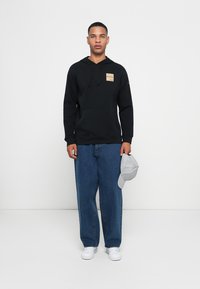 Felpa nera con cappuccio, tasca a marsupio frontale e patch del logo, abbinata a jeans di denim blu larghi e sneaker bianche. Tiene un cappellino grigio chiaro.