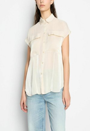 Camicia corta a maniche corte color beige chiaro con chiusura a bottoni, due tasche sul petto e una texture liscia, leggermente trasparente. Abbinata a jeans chiari.