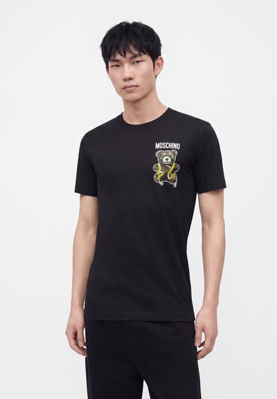 Νέος άνδρας που φοράει μαύρο t-shirt Moschino με γραφικό αρκουδάκι και κίτρινη ταινία στο στήθος, στέκεται μπροστά από ένα μονόχρωμο λευκό φόντο.