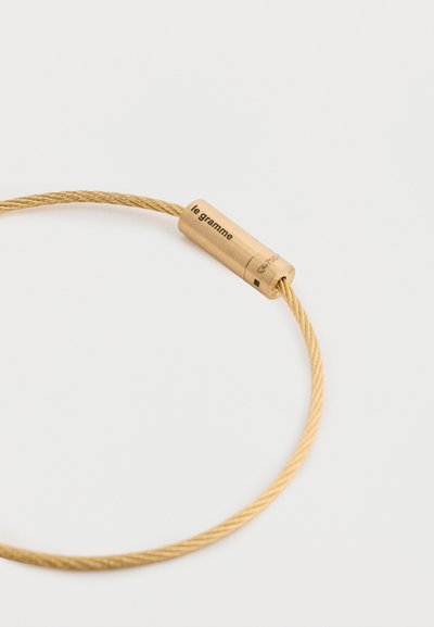 Χρυσό μεταλλικό βραχιόλι με κυλινδρικό κούμπωμα φέρει την ένδειξη "le gramme" και έχει υφή. Λείος, κομψός σχεδιασμός με διακριτική γυαλάδα.