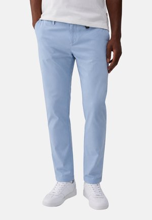 Uomo che indossa pantaloni skinny azzurri chiari e sneakers bianche, in piedi davanti a uno sfondo semplice con le mani lungo i fianchi.