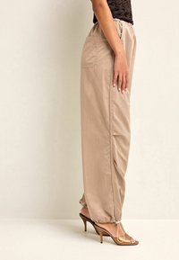 Beige snorebukser med sidelommer, der har en tapered benlængde og elastiske manchetter. Sammen med brune højhælede sandaler.