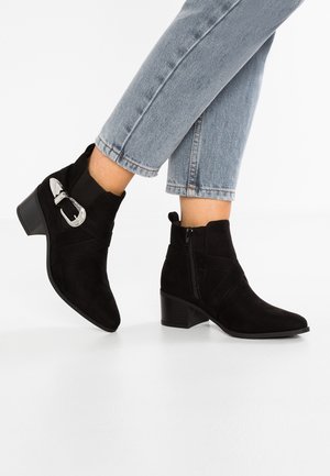 Bottes en suède noir montant à la cheville avec talons carrés, fermeture éclair latérale et boucle argentée décorative, portées avec un jean court bleu clair.