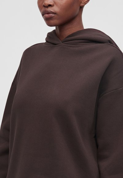 Personne portant un sweat à capuche marron foncé, visible des épaules au menton sur un fond clair et uni.