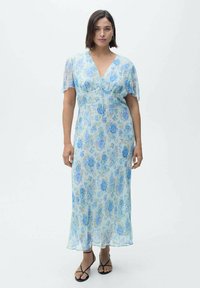 Langes, fließendes Kleid aus transparentem hellblauem Stoff mit floralem Muster. V-Ausschnitt und flatternde Ärmel. Kombiniert mit schwarzen Riemchensandalen.