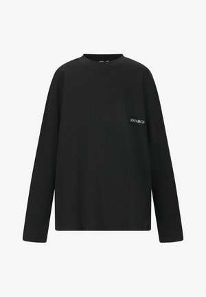 Sort langærmet skjorte, mørke shorts, hvide sokker og sorte sneakers. Modellen står med hænderne i lommerne mod en ensfarvet baggrund.