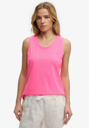 Femme portant un haut sans manches rose vif et un pantalon blanc à cordon, debout avec une expression neutre sur un fond uni.