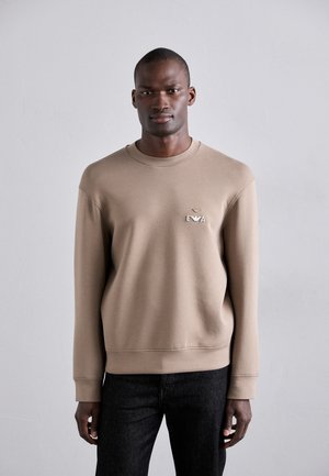 Light brown sweatshirt with a crew neckline and ribbed cuffs. Features an embroidered logo on the chest. Paired with dark jeans.