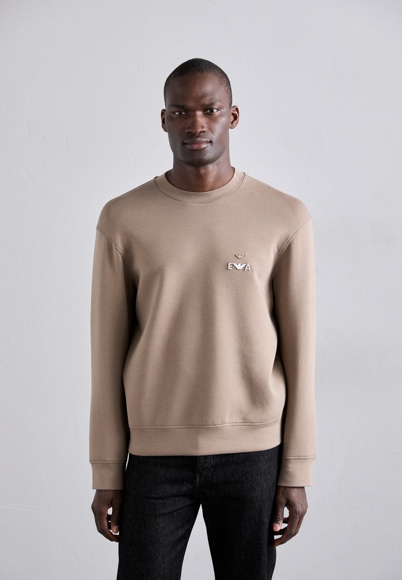 Lys brun sweatshirt med rund hals og ribbede mansjetter. Har et brodert logo på brystet. Kombinert med mørke jeans.