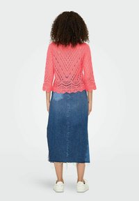 Haut en tricot ouvert rose avec des manches cloche 3/4, associé à une jupe midi en denim bleu avec un ourlet effiloché et une fente sur le côté, portée avec des baskets blanches.