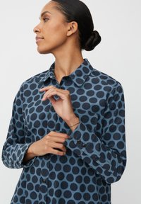 Camicia a maniche lunghe in tessuto blu navy con grandi pois blu scuro. Presenta un colletto classico, polsini con bottoni e taschino sul petto.