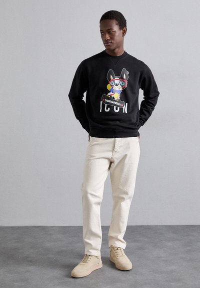 Black sweatshirt with a colorful graphic design, featuring a character with glasses, paired with cream-colored jeans and light tan shoes.