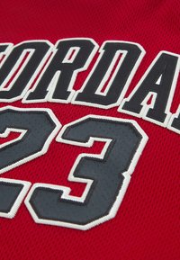 Sarkans acs tīkls džersijs ar izceltu melnu izšūtu tekstu "JORDAN" un numuru "23", apvilkti ar baltu kontūru, ar fakturētu virsmu.