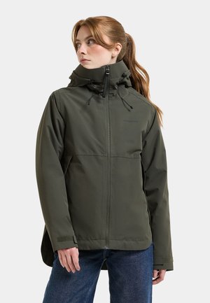 Vrouw met een donkergroene capuchontrui en blauwe spijkerbroek, kijkt opzij met haar haar in een paardenstaart.