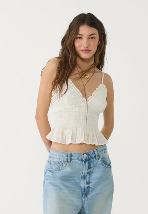 Top bianco a peplo con finitura testurizzata, spalline sottili e scollatura a onda. Abbinato a jeans high-waisted blu chiaro.