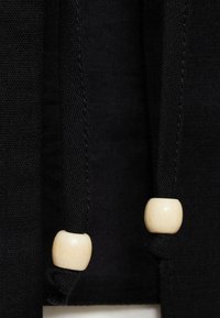 Mustapäällinen kangas, jossa on kaksi mustaa nyöriä, joista kummassakin on pyöreä, sileä puuhelmi akcenttina. Kankaan pinta näyttää kudotulta ja tiheältä.