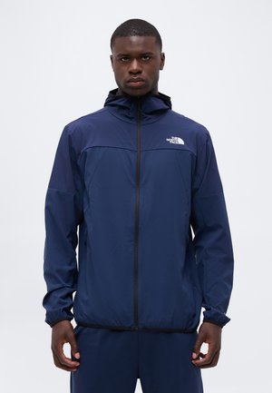 Jeune homme portant une veste à capuche The North Face bleu marine zippée, debout devant un fond gris clair uni.