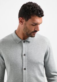 Lichtgrijs gebreid polo-shirt met een omgeslagen kraag, voorzien van drie bruine knopen en een zachte, gestructureerde stof.