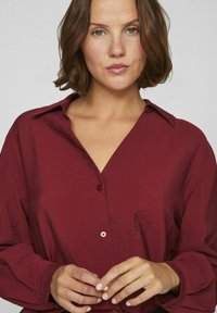 Camisa burdeos de botones con una tela de textura fina, mangas largas, cuello con muesca y un detalle de un solo botón en el cuello.