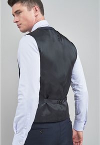 Gilet noir en satin pour homme avec une ceinture ajustable à l'arrière, porté sur une chemise blanche avec des accents bleus et un pantalon noir. Présente un design élégant et une texture lisse.