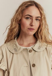 Beige trenchcoat med en rund krave, broderet blomstermotiv, to brune knapper og indsamlede ærmer, båret over en hvid top.