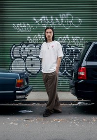 Fehér rövid ujjú póló rózsaszín grafikával, barna csíkos nadrág, fekete cipő, két autó között állva, zöld háttéren graffiti.