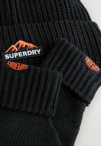 Zwarte gebreide beanie met een ribstructuur. Voorzien van een oranje en witte "SUPERDRY" logo patch met een bergontwerp.