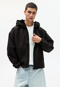 Schwarze Kapuzenjacke mit Reißverschluss, elastischen Bündchen und glattem, wasserdichtem Material, kombiniert mit einem weißen T-Shirt und hellblauen Jeans.