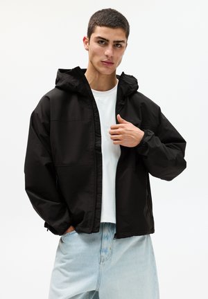 Veste à capuche noire avec fermeture éclair à l'avant, poignets élastiques et un tissu imperméable lisse, associée à un t-shirt blanc et un jean bleu clair.