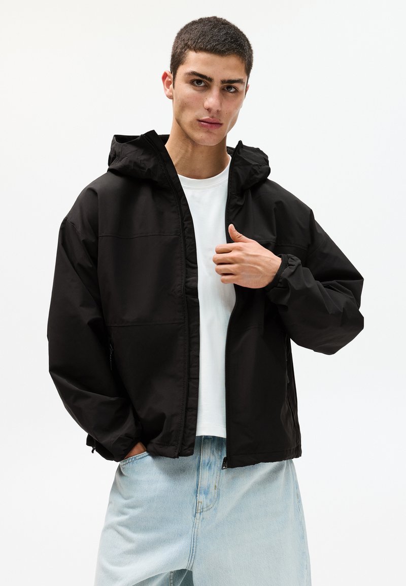 Schwarze Kapuzenjacke mit Reißverschluss, elastischen Bündchen und glattem, wasserdichtem Material, kombiniert mit einem weißen T-Shirt und hellblauen Jeans.