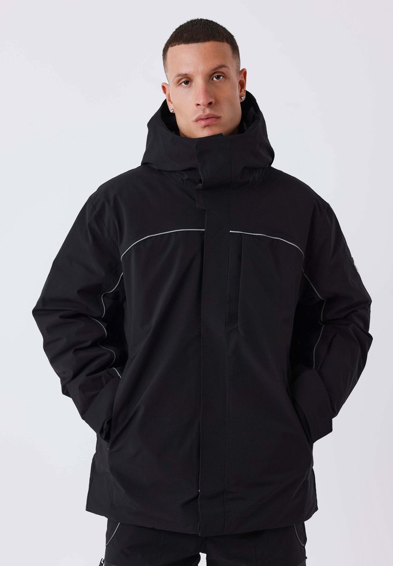 Zwarte waterdichte jas met een hoge kraag, ritssluiting en reflecterende accenten langs de schouders. Heeft een gladde textuur en een oversized pasvorm.