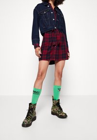 Denim cropped jack over een rood en zwart geblokt shirtjurk. Groene kniesokken met zwarte tekst, gecombineerd met bloemenpatroon zwarte laarzen.