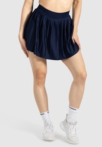Marineblauwe geplooide rok met een brede tailleband, gemaakt van een gladde stof. Gedragen met witte sportieve schoenen en sokken met zwarte branding.