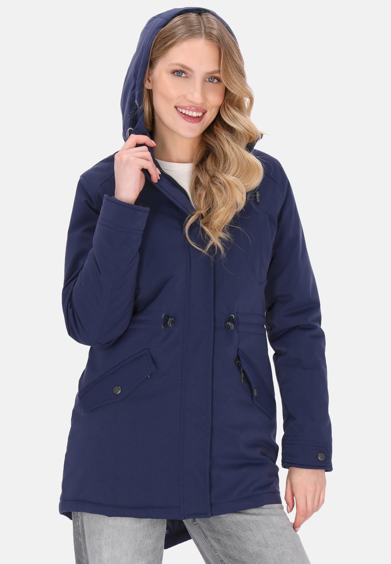 Kobieta uśmiechająca się, nosząca granatową kurtkę z kapturem, z zamkiem błyskawicznym i kieszeniami na napy, trzymająca kaptur jedną ręką.