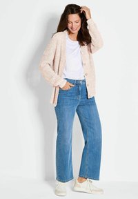 Helles rosa strukturiertes Cardigan mit Knöpfen über einem weißen T-Shirt, kombiniert mit blauen weit geschnittenen Jeans und weißen Sneakers.