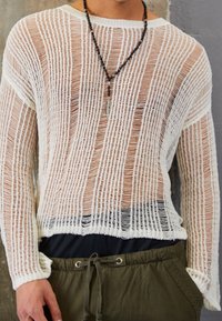 Person som bär en löst stickad vit långärmad tröja över en svart t-shirt, olivgröna byxor med snodd och ett svart pärlhalsband med en hänge.