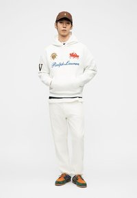 Jonge man met een witte Ralph Lauren hoodie, witte broek, een bruine pet en meerkleurige veter schoenen, staand met zijn handen in zijn zakken.