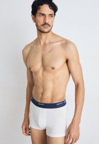 Άνδρας χωρίς πουκάμισο με κοντά, σγουρά μαλλιά, φοράει άσπρο μποξεράκι με σκούρο μπλε λάστιχο μέσης GANT, στέκεται μπροστά σε ένα απλό φωτεινό φόντο.