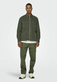 Chaqueta de pana verde oliva con dos bolsillos frontales, cierre de cremallera y pantalones a juego de color verde oliva. Lleva puesta una camiseta blanca y zapatillas claras.