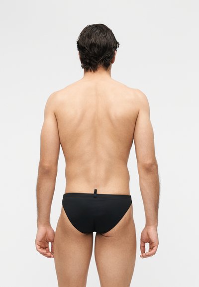 Άνδρας με κοντά σκούρα μαλλιά, που στέκεται γυρισμένος προς τα πίσω, φορώντας μαύρα ανδρικά μαγιό σε απλό ανοιχτό φόντο.