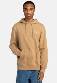 Tan cotton hoodie with drawstring hood, front pocket, ribbed cuffs, and hem; features white logo text on the chest. Styled with blue jeans.