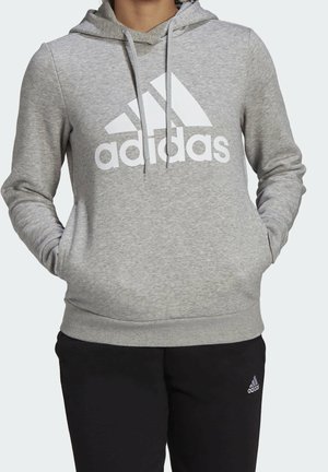 Person iført en grå Adidas-hættetrøje med stort hvidt logo og sorte Adidas-bukser, hænderne i hættetrøjelommerne.