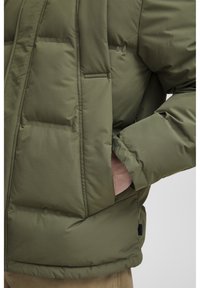 Olive green puffer jacket with a quilted design, featuring side pockets and a smooth texture. The hem is slightly longer in the back.